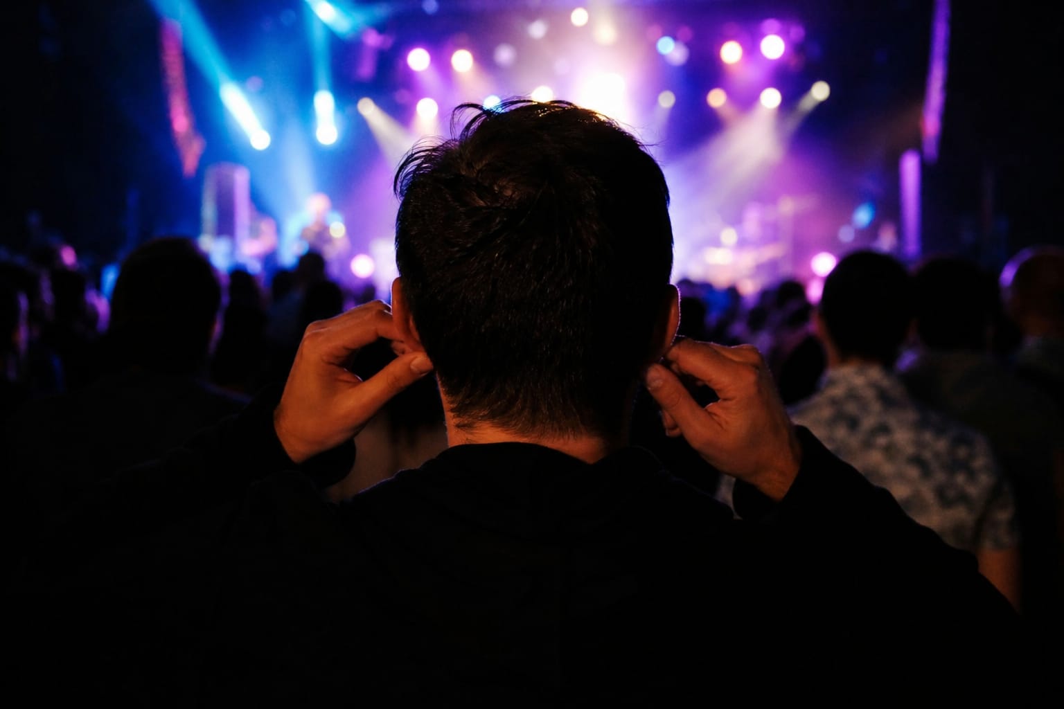 Personne se bouchant légèrement les oreilles dans une ambiance de concert, lumières colorées en arrière-plan