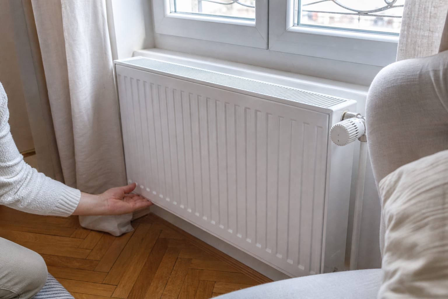 Main posée sur le bas d'un radiateur à eau blanc installé sous une fenêtre, avec sa tête thermostatique visible sur le côté